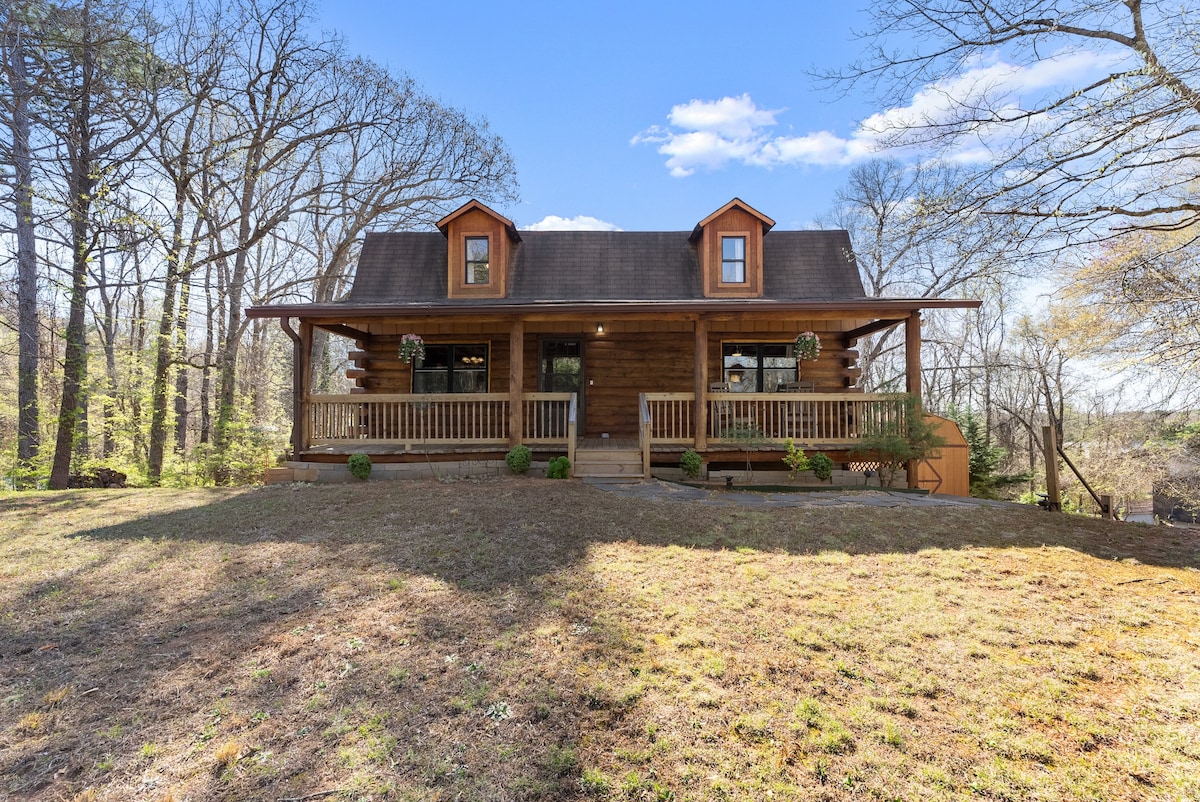 A log cabin is set amidst a natural landscape, featuring a welcoming front porch with a sturdy railing. Symmetrical windows are adorned with hanging flower baskets, allowing natural light to fill the interior. The surrounding grassy area is framed by trees and open sky.