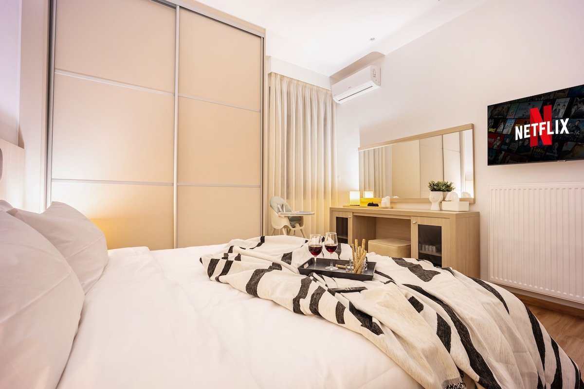 A cozy bedroom is presented, featuring a spacious bed draped with a striped blanket. A modern dresser is positioned beside the bed, complemented by a wall-mounted TV displaying the Netflix logo. Soft lighting enhances the inviting atmosphere, while a large closet adds functionality.