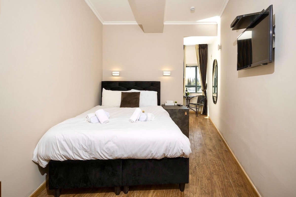 The spacious room features a large bed with soft linens. A small seating area and a wall-mounted TV are also visible. Natural light enters through a large window, revealing views of the surrounding area.