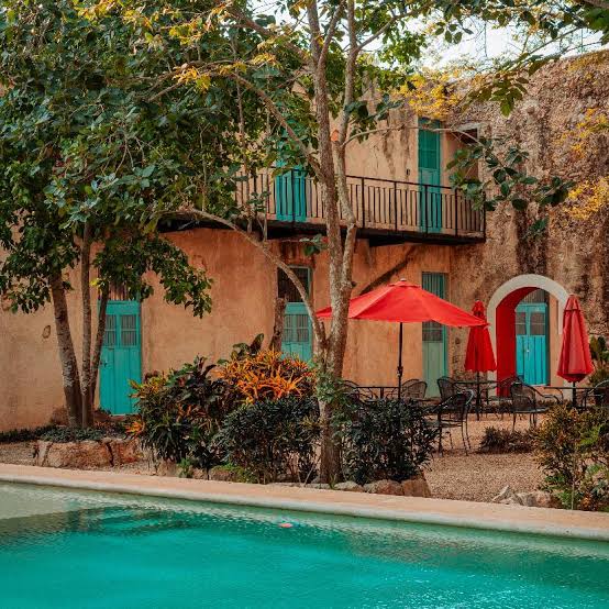 A rustic outdoor area features a calm swimming pool surrounded by colorful tropical vegetation. Bright red umbrellas provide shade over several seating arrangements. The building's weathered walls complement the teal doors, adding charm to the peaceful ambiance.
