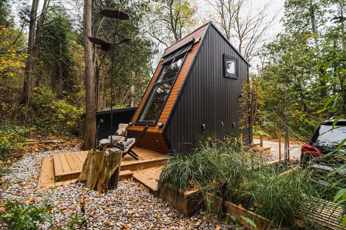 The tiny home is surrounded by trees and gravel pathways, featuring a sloped roof and large angled windows. A wooden deck extends in front, blending with the natural landscape. Soft grasses and rustic stones are present, emphasizing the serene outdoor setting.