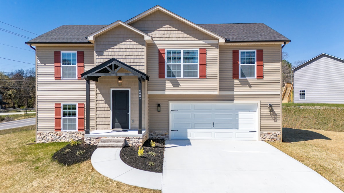 The exterior of the new construction home showcases a welcoming entrance with a covered porch and an inviting front door. Red shutters accentuate the beige siding, while a neatly maintained lawn and driveway lead to a spacious two-car garage.