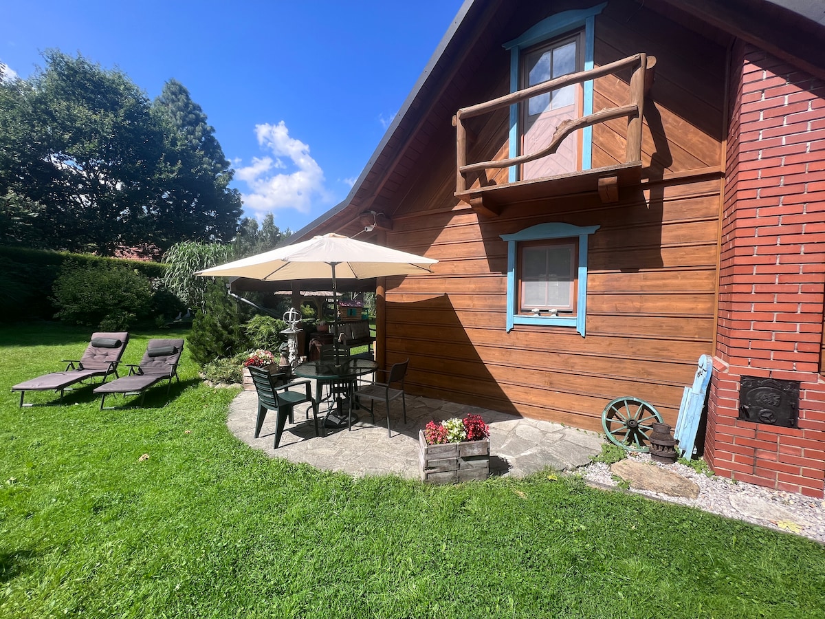 A charming outdoor patio area features a shaded table with chairs under a large umbrella. Two lounge chairs are placed on the grassy lawn, surrounded by well-kept flowerbeds. The wooden cabin offers a rustic appeal with blue window frames and a balcony overlooking the garden.