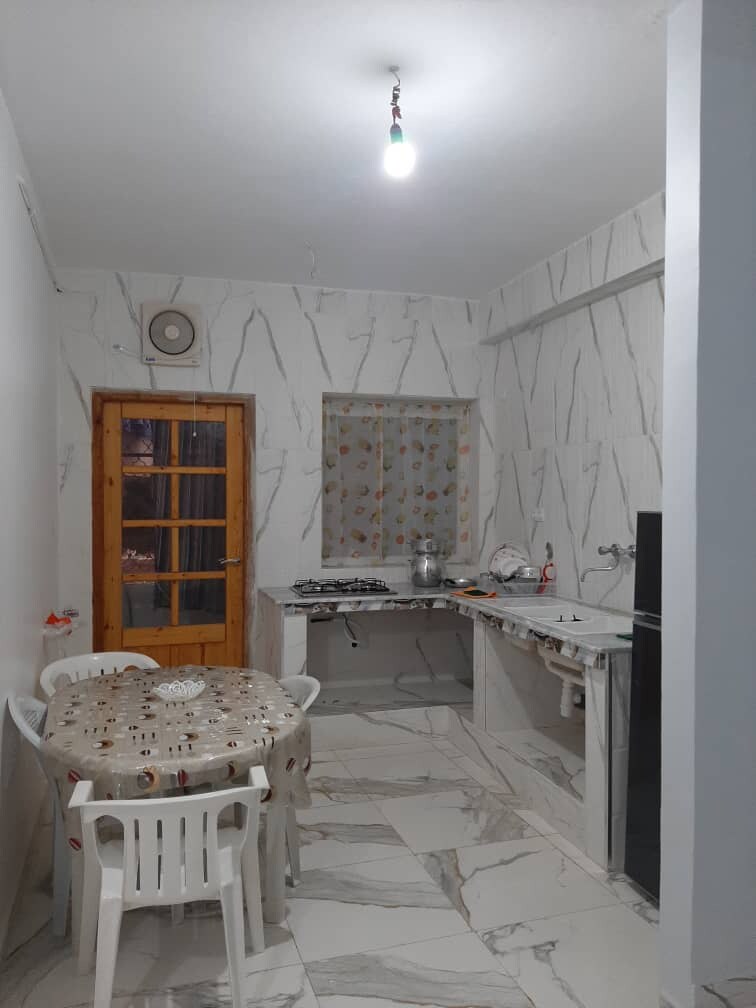 A kitchen area features light-colored marble walls and floor tiles. A round table with a decorative tablecloth is surrounded by four plastic chairs. A window with a sheer curtain allows natural light, and gas burners are visible on the countertop, demonstrating functionality.