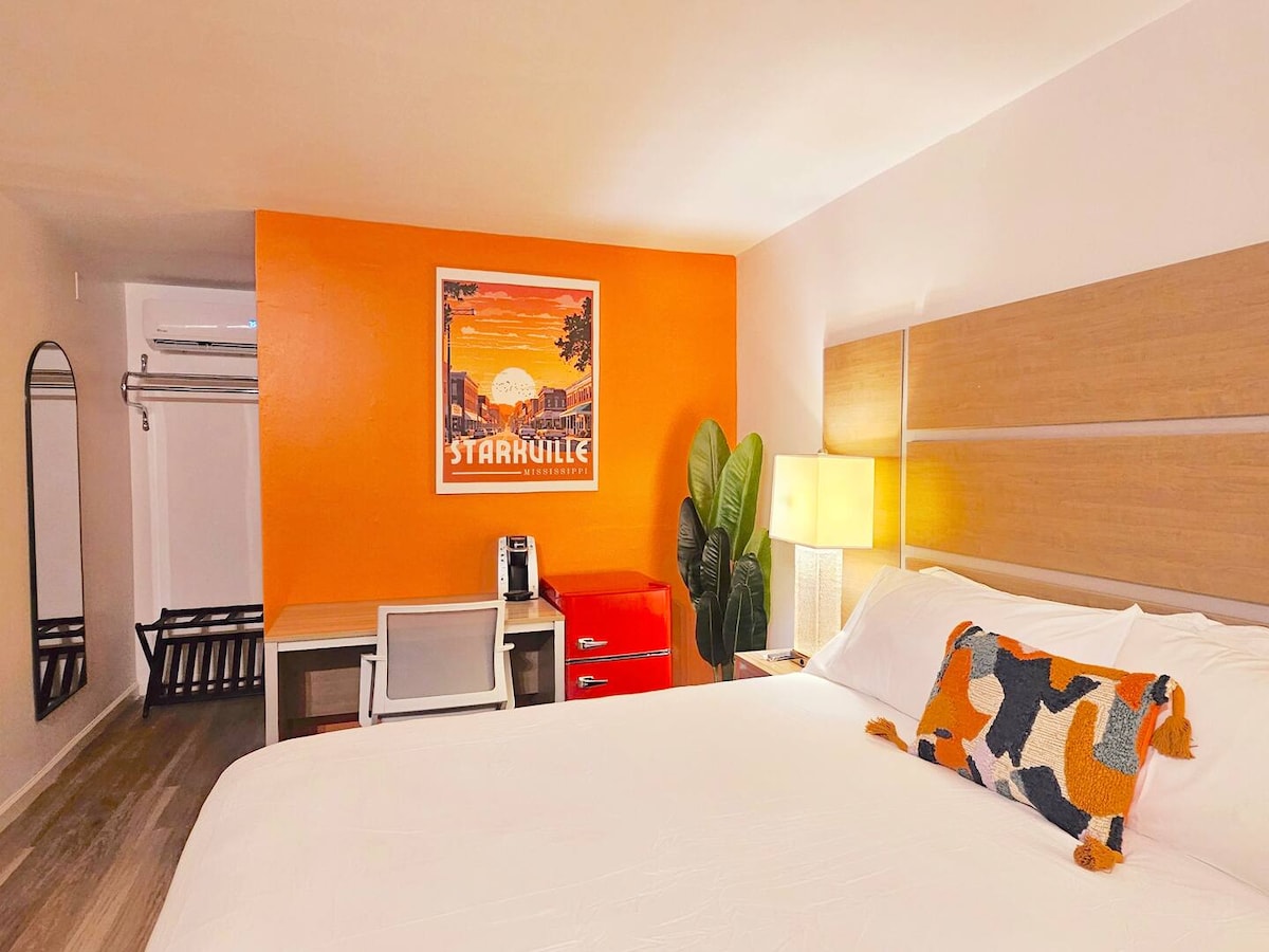A bright sleeping area features an inviting orange accent wall. A spacious bed is adorned with a patterned throw pillow. A compact workspace with a chair is positioned adjacent to the bed, while a modern lamp and a green plant add warmth to the room.