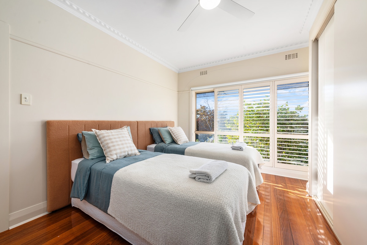 The room features two twin beds, each with textured blankets and neatly arranged pillows. Natural light floods the space through large windows adorned with wooden blinds, while polished wooden floors add warmth to the environment.