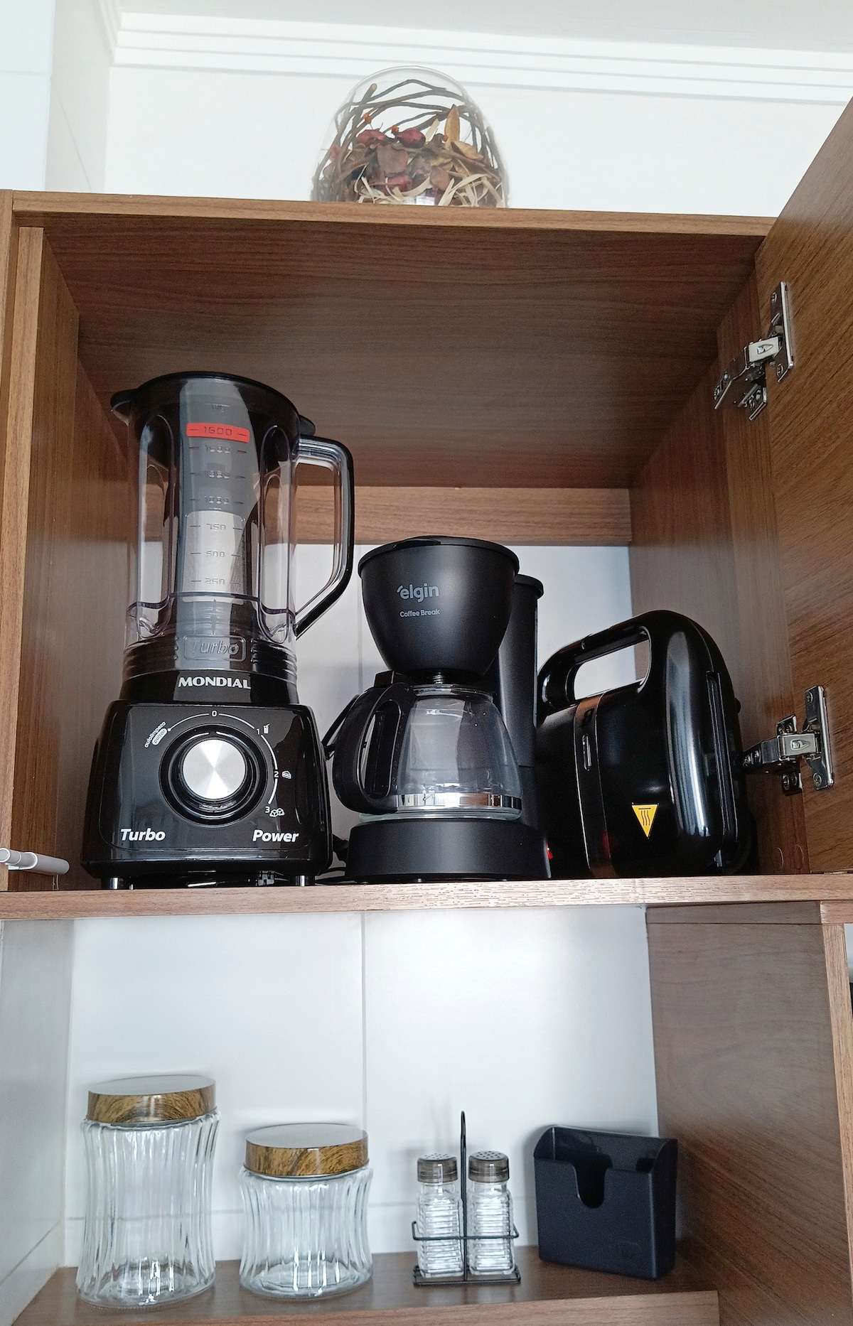 A kitchen cabinet shelf displays essential appliances. A blender, coffee maker, and toaster are neatly arranged, while two glass jars and a spice container are positioned below. The wood grain of the cabinet adds warmth to the kitchen setting.