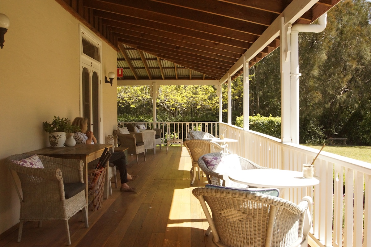 A spacious verandah features a wooden floor and a covered ceiling supported by beams. Comfortable seating is arranged around tables, with potted plants adding a touch of greenery. Natural light filters in, creating an inviting space for relaxation.