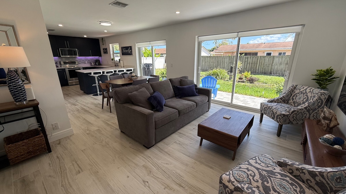 An open living area features a comfortable seating arrangement with gray sofas and patterned armchairs. Large windows allow natural light to fill the space, offering a view of the outdoor area. A modern kitchen is visible in the background, equipped with sleek appliances and bar seating.