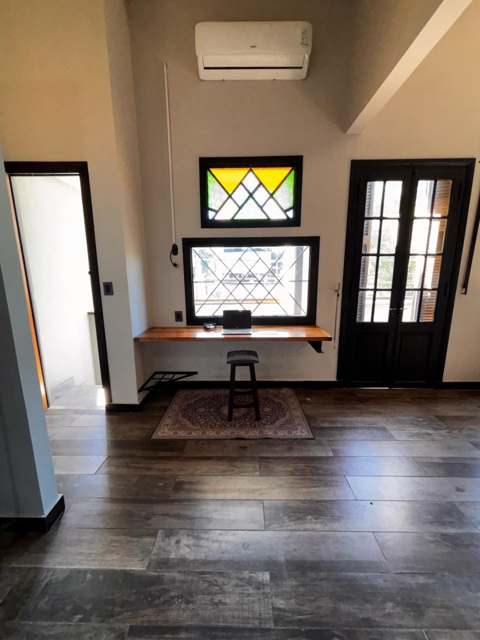 A workspace features a wooden desk positioned under a colorful stained glass window, providing natural light. A small stool is placed in front of the desk, and a decorative rug lies on the floor. The room showcases wooden flooring and a doorway leading to another space.