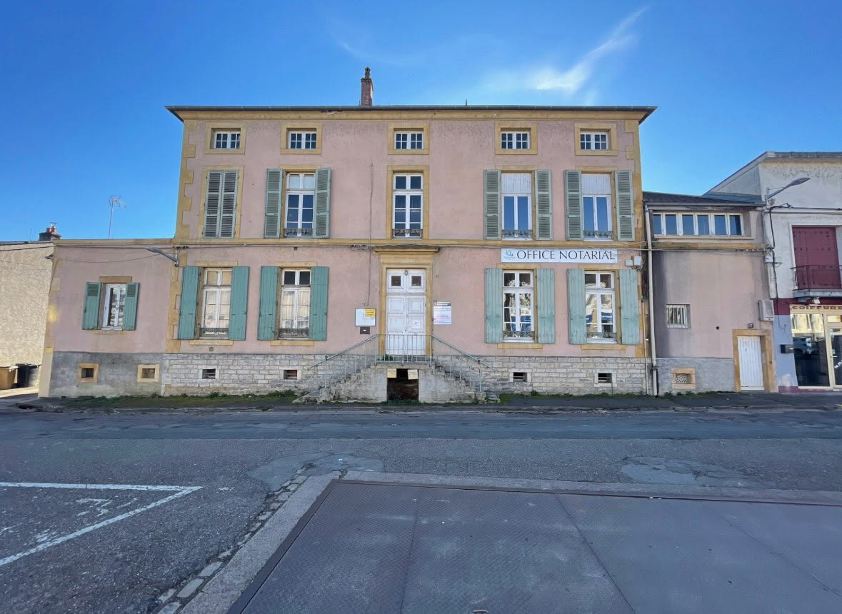 The image displays a two-story building with a pink facade and green shutters. A central set of stairs leads to the double entrance doors, flanked by large windows. The surrounding area includes a parking lot, with clear skies overhead.
