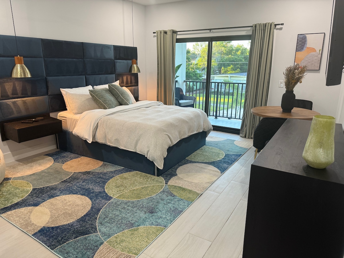 A spacious sleeping area features a modern bed with a plush headboard and neutral bedding. A round dining table is present nearby, and a large sliding door provides access to a private balcony. Soft, circular rugs add texture to the light-toned floor.