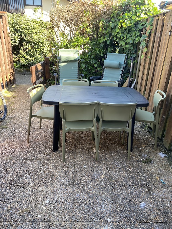 An outdoor dining area is presented with a rectangular table surrounded by six stackable chairs. Folding lounge chairs are positioned nearby, against a backdrop of greenery and fencing. The ground is made of a gravel surface, providing a natural setting.