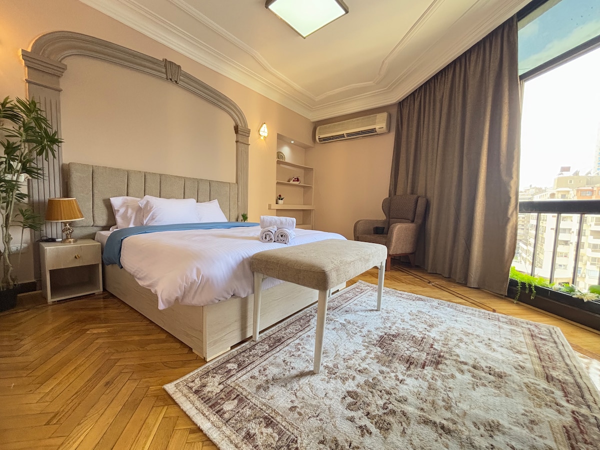 A spacious bedroom features a light-colored bed adorned with fresh linens and folded towels. A bench sits at the foot of the bed. Large windows allow natural light to fill the room, and a comfortable chair is positioned nearby, with soft curtains adding warmth to the space.