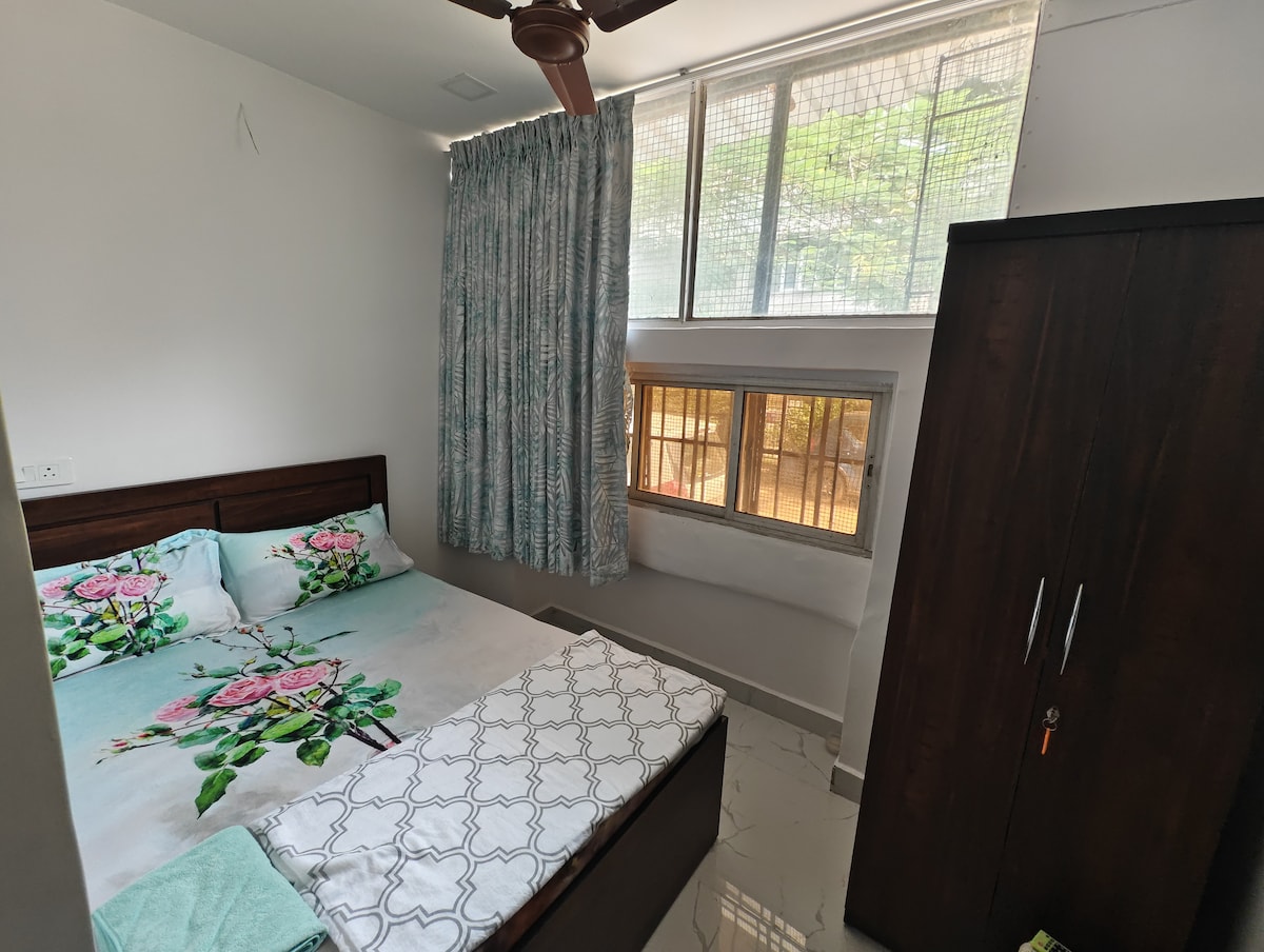 A well-appointed room features a comfortable bed with floral-patterned bedding. Natural light filters through large windows, complemented by light-filtering curtains. A wooden wardrobe stands nearby, contributing to the modern decor. The flooring is sleek, enhancing the overall freshness of the space.