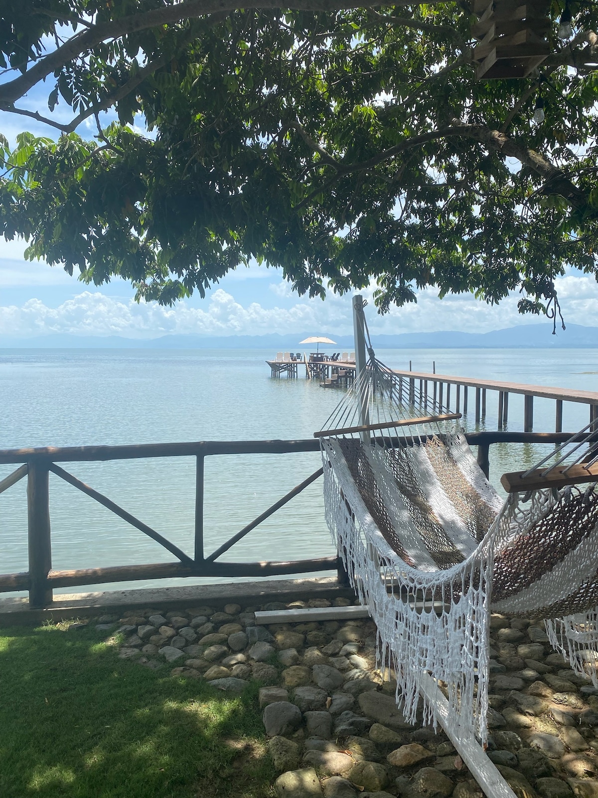 Two hammocks are gently suspended between posts, surrounded by lush greenery. A view of a tranquil water body is visible in the background, with a wooden dock extending into the water, creating a serene outdoor space for relaxation.