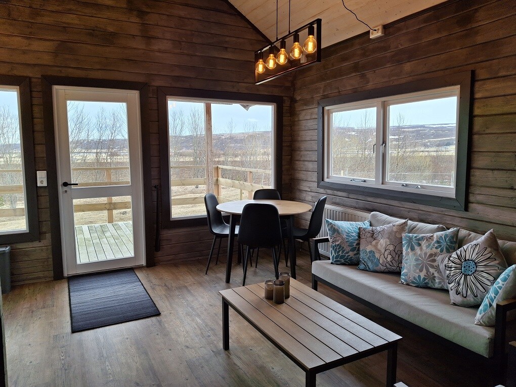 A living area is showcased with large windows allowing natural light to illuminate the space. A circular dining table with black chairs is positioned nearby. A cozy sofa is adorned with decorative pillows, and a wooden coffee table is placed in front, enhancing the inviting atmosphere.