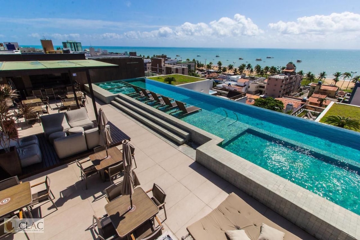 An elevated pool area features sleek lounge chairs alongside a stunning infinity pool overlooking the ocean. The clear blue water blends into the horizon, while shaded seating areas invite relaxation. The surrounding rooftop is lined with palm trees, creating a serene atmosphere.