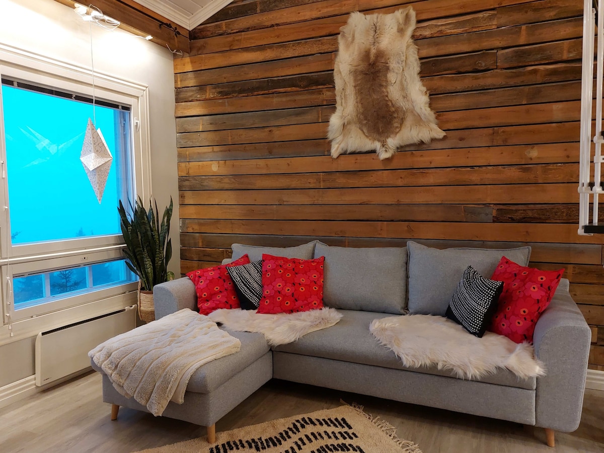 A comfortable gray sofa with a sectional design is complemented by vibrant red and black decorative pillows and cozy throws. The backdrop features a textured wood panel wall, while a large window allows natural light to brighten the space. A potted plant adds a touch of greenery.