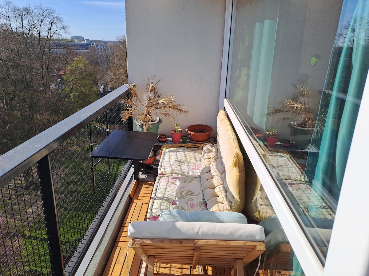 A small outdoor balcony is shown, featuring a wooden chair and a cushioned bench. Potted plants are arranged on a small table and along the railing. Glass doors provide a view of the surrounding greenery in the background.