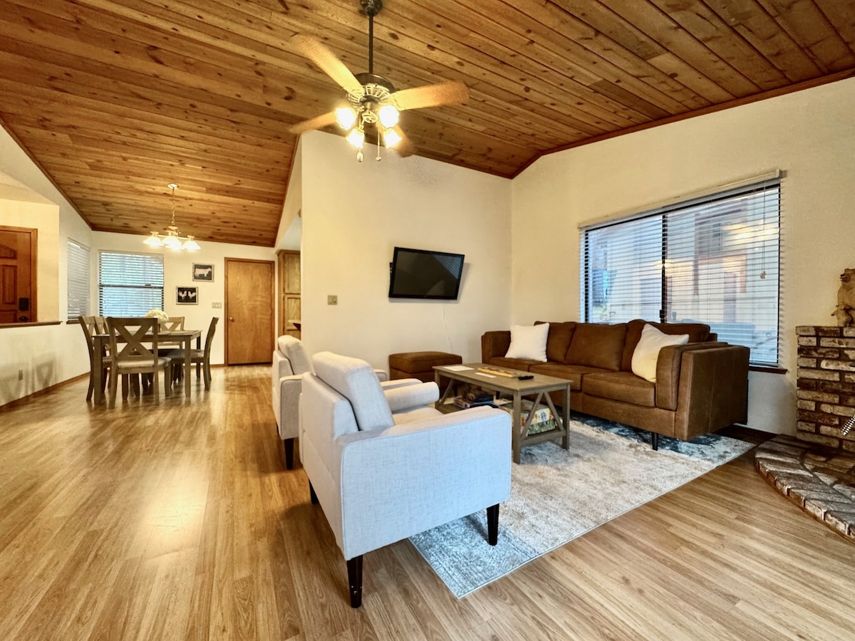 The living area features a spacious layout with comfortable seating arranged around a coffee table on a soft area rug. Large windows allow natural light to fill the room, complemented by warm wooden ceiling panels. A television mounted on the wall enhances the inviting atmosphere.