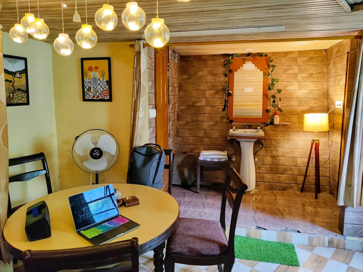 A dining area features a round table with four chairs, thoughtful lighting provided by hanging glass pendant lights. A fan is positioned nearby. In the background, a mirror is framed by natural wood, and a lamp stands beside a small sink area, enhancing the room's functionality.
