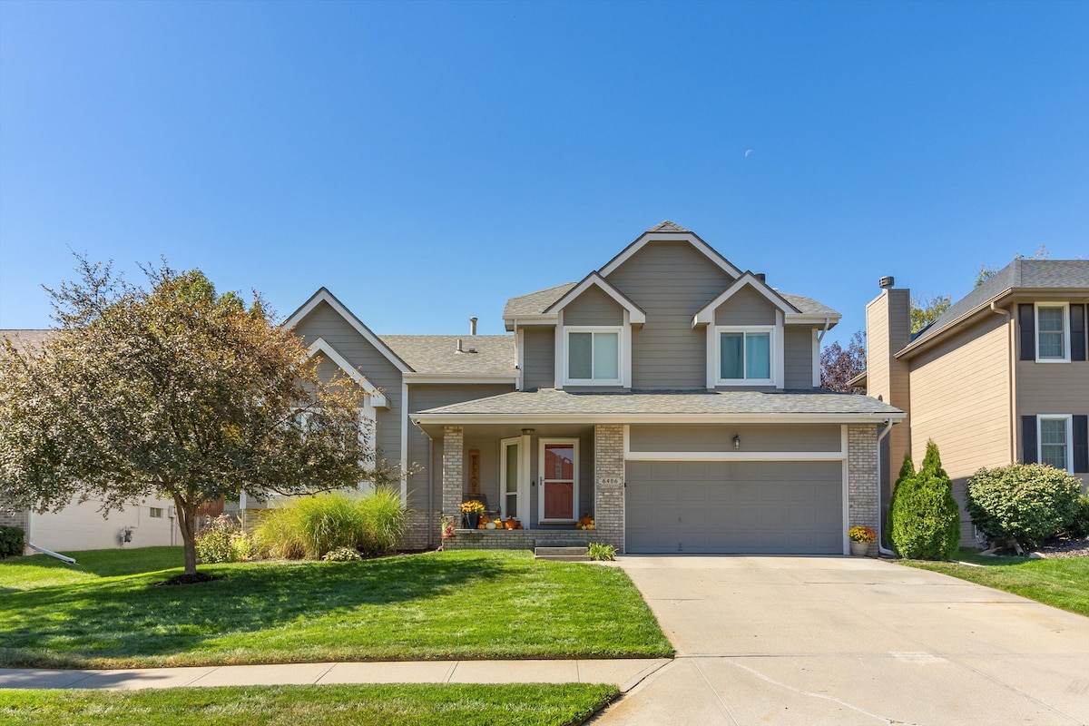 The exterior of a two-story house is featured, showcasing a warm grey facade with stone accents. A well-kept lawn surrounds the property, complemented by a paved driveway leading to a two-car garage. The entrance is framed by autumnal plants, enhancing the welcoming appeal.