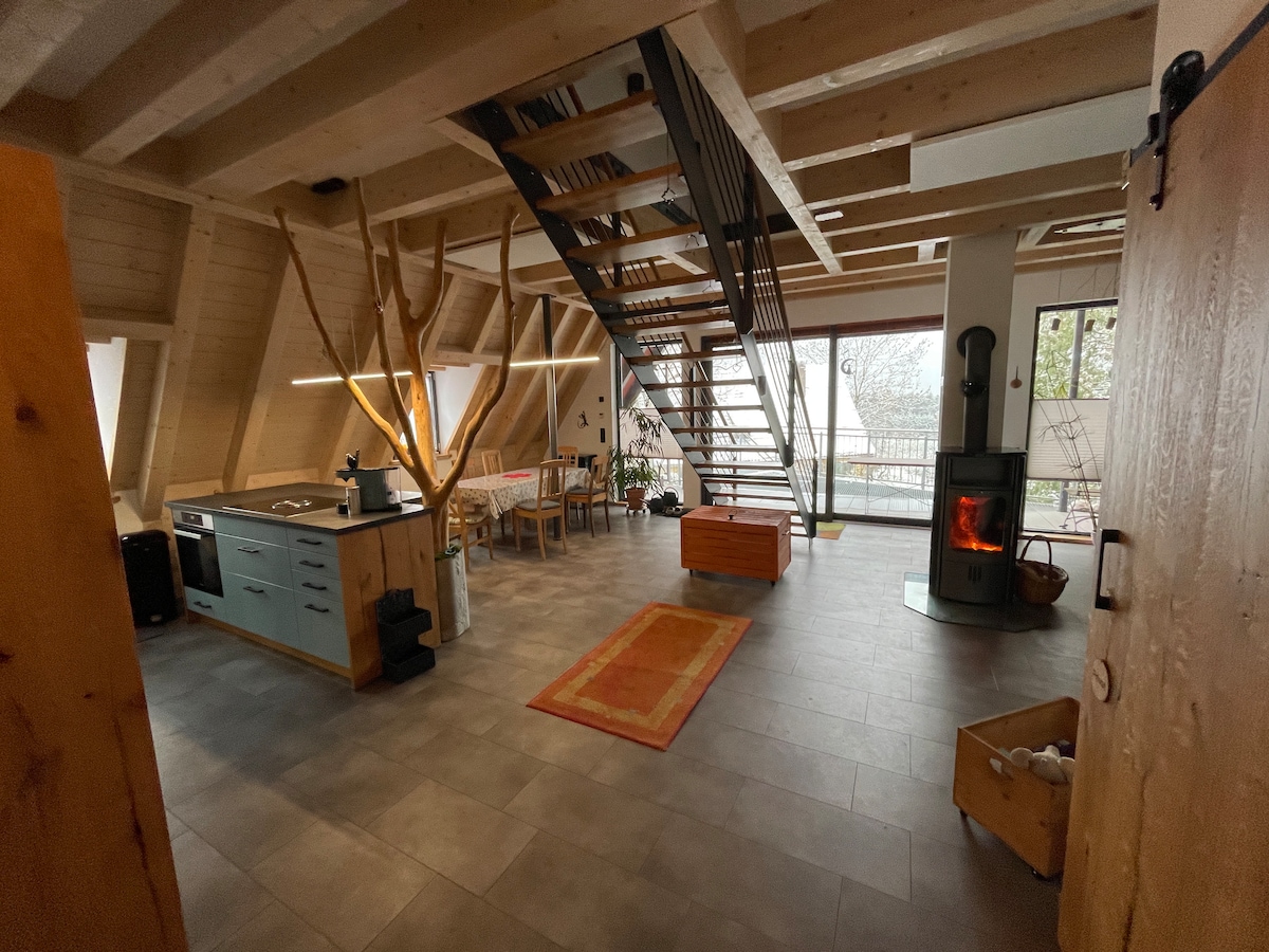 An open-plan living area showcases a modern kitchen with a wooden island, while a dining space is positioned nearby. A staircase leads to the upper level. Natural light floods the room through large glass windows, complemented by a corner fireplace and warm wooden elements.