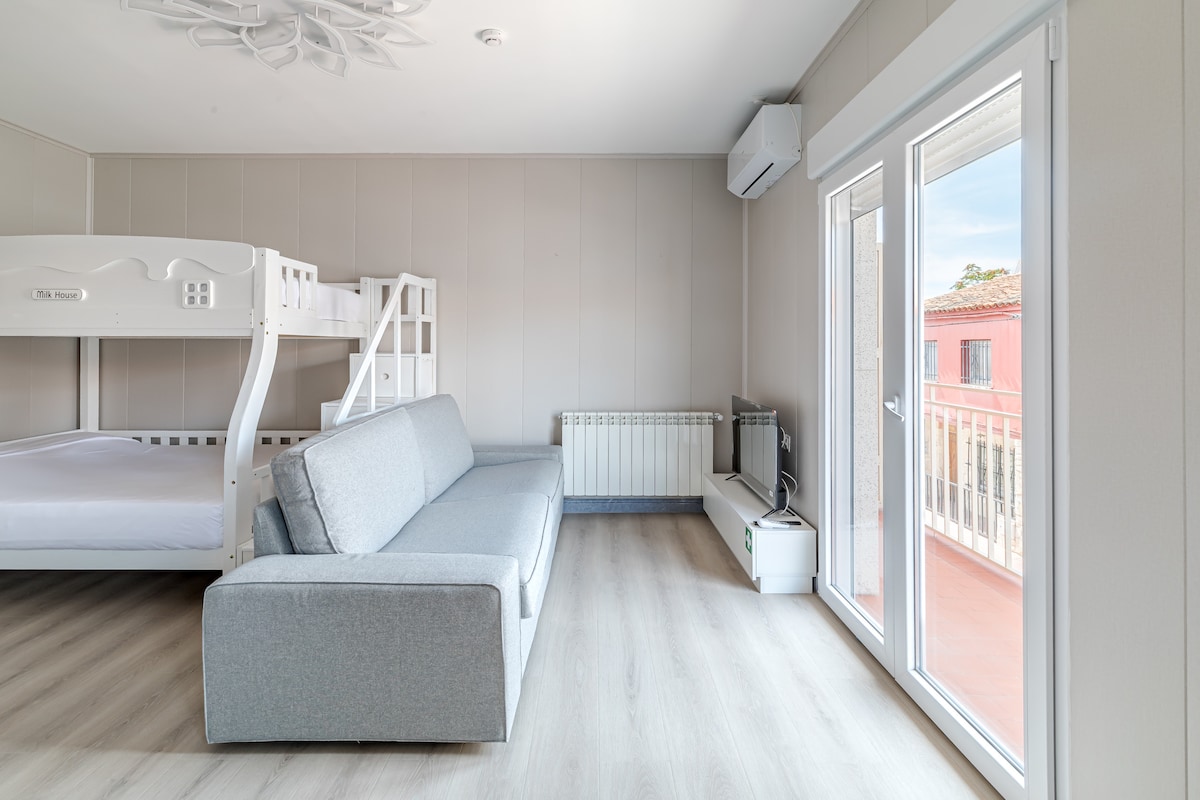 A bright and airy living space features a light color palette. A comfortable grey sofa is positioned across from a bunk bed, with the bottom bunk accommodating a spacious mattress. Large sliding doors provide natural light and open to a small balcony area.