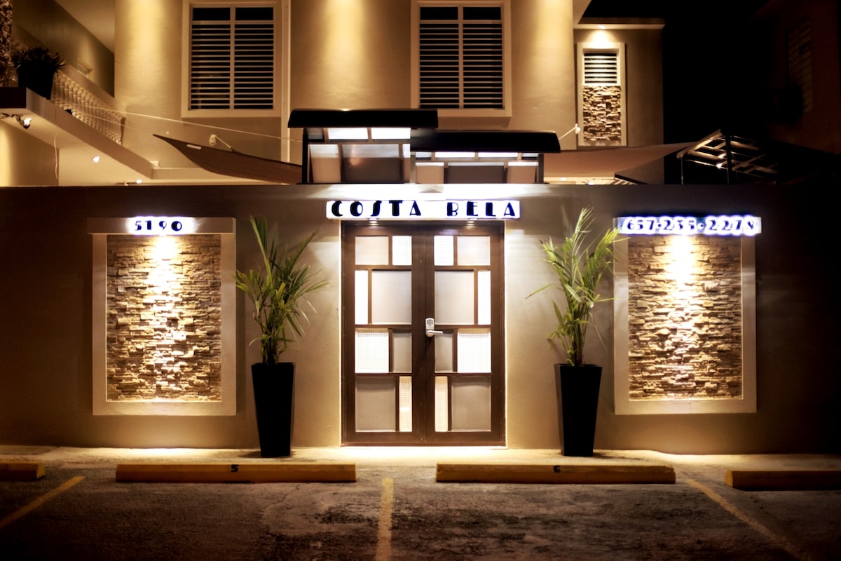 The entrance to Costa Bela is showcased, featuring a large wooden door framed by decorative stone walls and illuminated signage. Potted plants stand on either side of the door, complementing the welcoming atmosphere of the well-lit exterior.