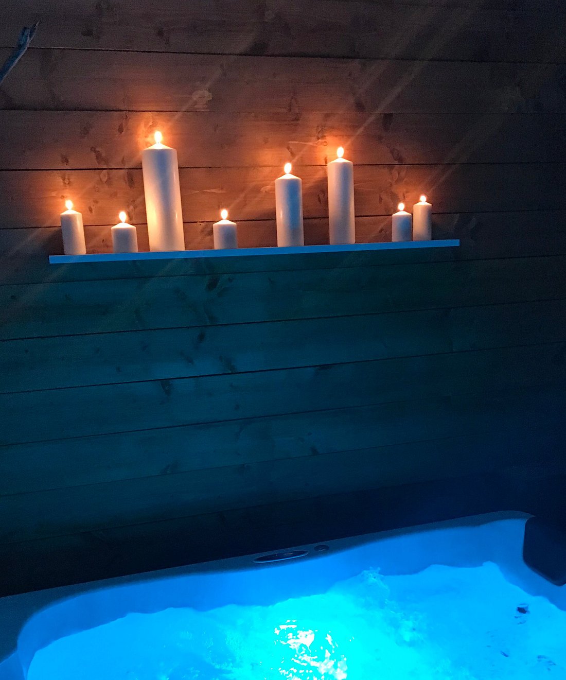 A serene spa area is illuminated by candles placed on a shelf against wooden paneling. Soft blue lighting emanates from the spa, creating a calming ambiance that invites relaxation.