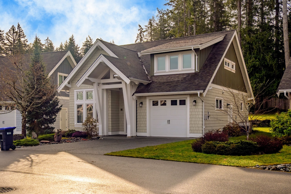 A charming two-story home is set amidst greenery, featuring a gabled roof and a welcoming entrance. Large windows allow natural light to flow in, while manicured landscaping complements the facade. A paved driveway provides access, creating an inviting ambiance.