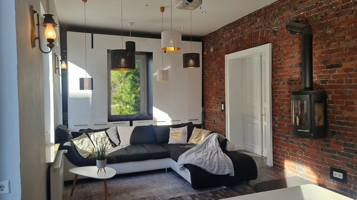 A modern living area features a sectional sofa adorned with decorative pillows and a soft throw blanket. A sleek coffee table sits in front, with light filtering through a large window. Exposed brick walls add character, while stylish pendant lights hang from the ceiling.