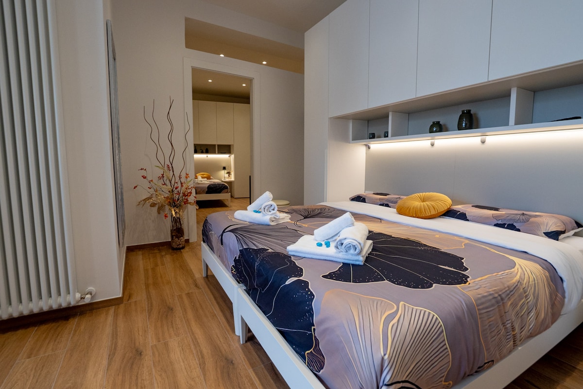 A well-appointed bedroom is presented with a spacious bed featuring floral-patterned bedding. Soft lighting enhances the room's features, including a set of towels neatly placed at the foot of the bed and decorative elements placed nearby. A glimpse of the en-suite bathroom is visible in the background.