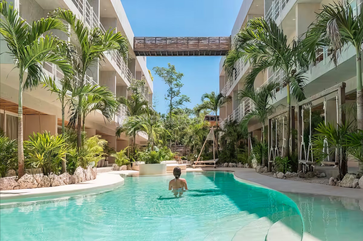 A tranquil pool area is surrounded by lush greenery and elegant buildings. Palm trees frame the water, where a person is seen relaxing. A bridge connects the upper levels of the structure, enhancing the open and inviting atmosphere of the space.