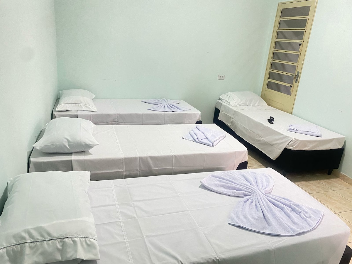 A bright, simple guest room features three individual beds, each covered with white linens and accompanied by fresh towels. Light green walls create a calm setting, while a door with frosted glass adds natural light. Neutral-colored floors complement the overall space.