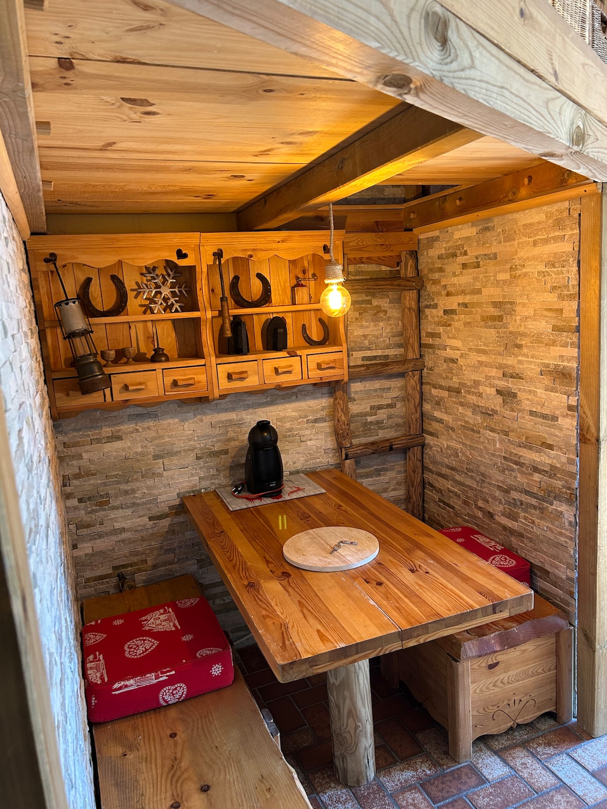 A rustic dining area features a wooden table set against a stone wall. Shelves above display assorted items, including a decorative piece and black utensils. Two red upholstered seats are positioned on either side of the table, with warm lighting illuminating the space from a hanging bulb.
