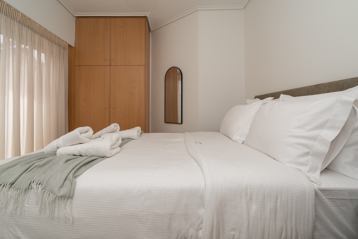 A spacious bedroom features a comfortable bed with white linens and neatly arranged towels. Light is gently filtered through sheer curtains, illuminating the room. A modern mirror and wooden wardrobe add functional elements, contributing to a welcoming atmosphere.