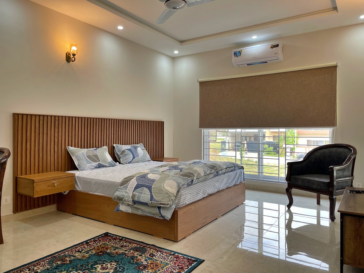 A bright guest room featuring a spacious bed with a patterned quilt, complemented by an armchair and window with light-filtering shades. A wooden accent wall adds texture, while polished flooring reflects natural light, creating a comfortable and well-appointed space for relaxation.