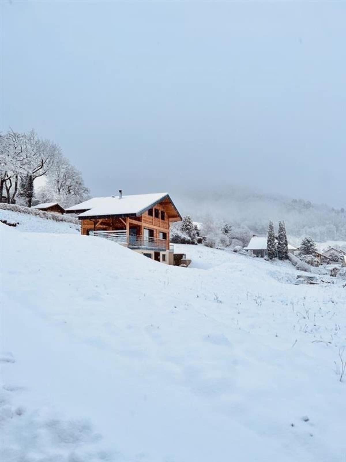 A recently constructed chalet is visible amidst a serene snowy landscape. The wooden exterior and multiple stories are highlighted against the soft white blanket of snow. Surrounding trees are lightly dusted, contributing to the tranquil winter scene.