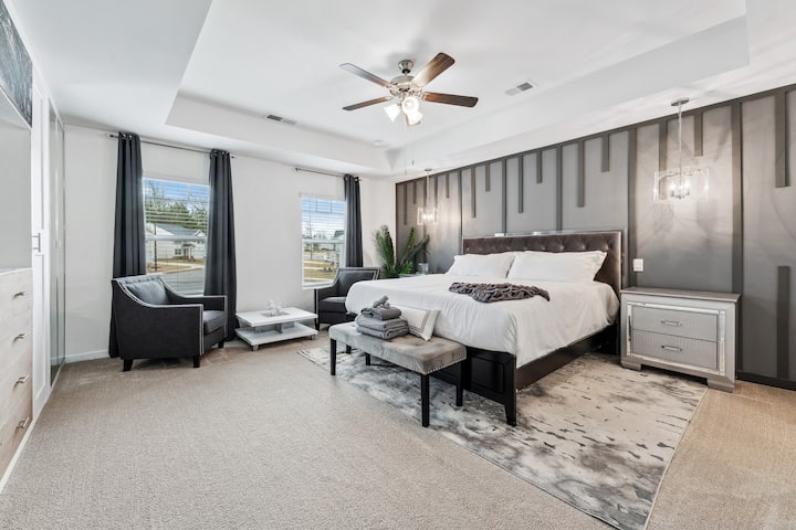This sophisticated primary bedroom features a king bed with a statement accent wall, pendant lighting, and a tray ceiling with fan. Relax in the cozy sitting area by the window or unwind with the serene, modern decor throughout.