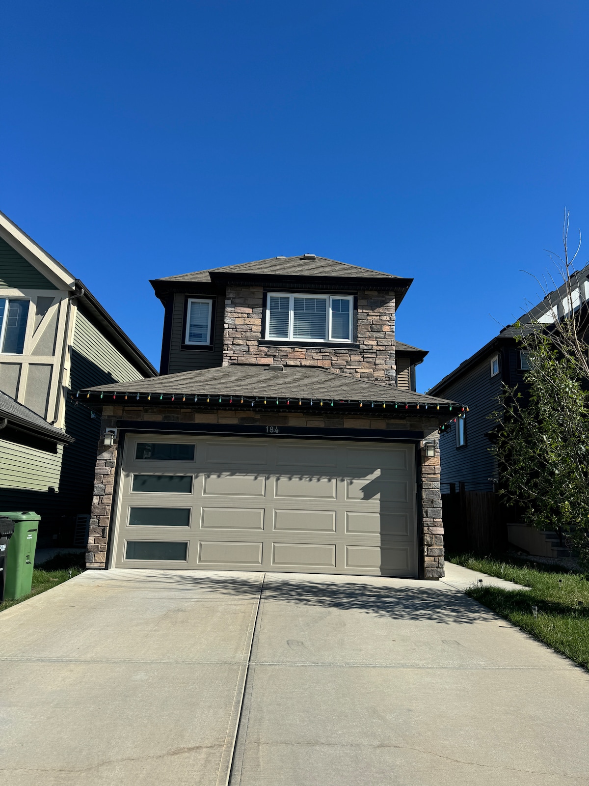 The exterior of the vacation home showcases a two-story structure with a stone facade and a three-car garage. A well-maintained driveway leads to the garage, bordered by greenery and residential architecture. The clear blue sky above enhances the inviting appearance of the home.