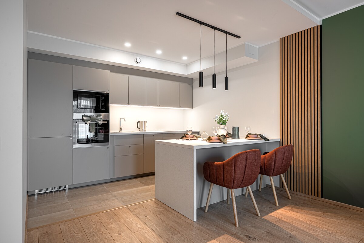 A modern kitchen is highlighted with sleek cabinetry and minimalist design. A spacious island serves as a dining area with two upholstered chairs. Soft, overhead lighting illuminates the counter, while large windows enhance the overall brightness of the space.