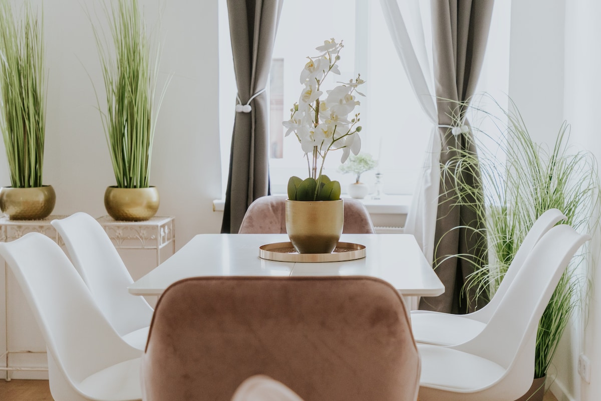 A dining area features a sleek white table surrounded by four contemporary chairs. A decorative arrangement of greenery and an orchid centerpiece is placed on the table, while two tall plants are positioned in golden pots on either side of the window, which is framed by soft curtains.