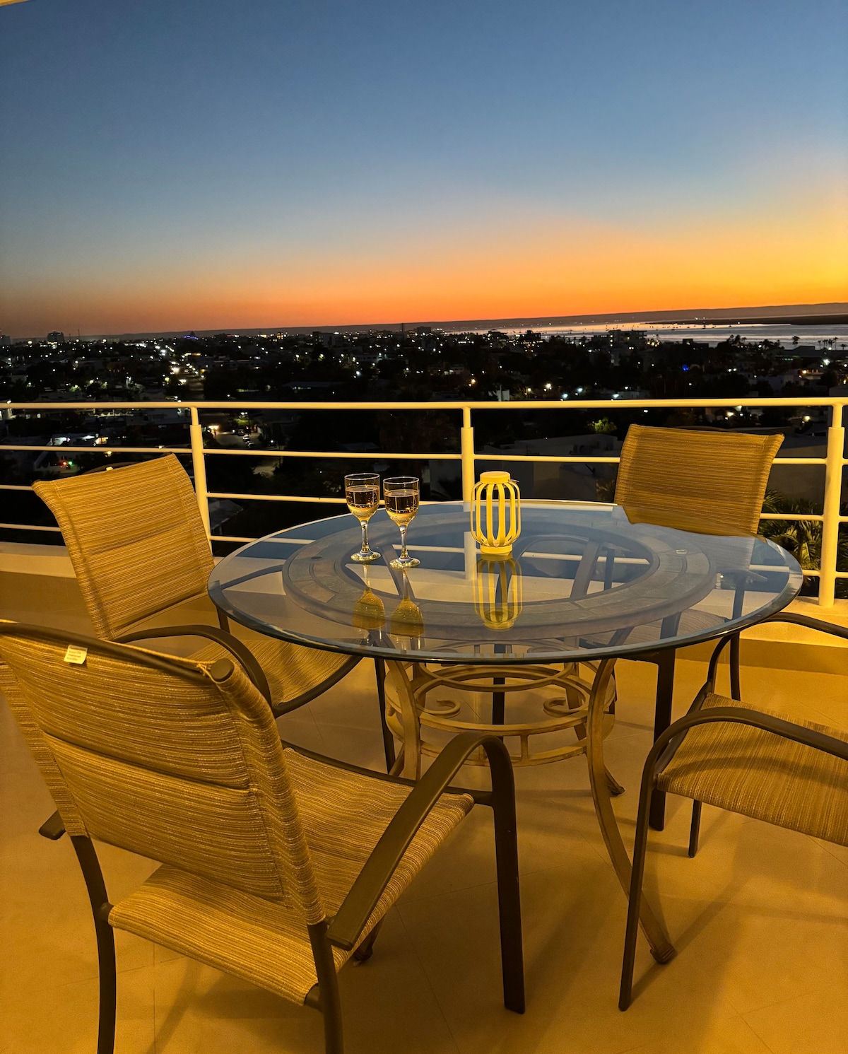 A round glass table is set with two glasses and a decorative item, placed on a balcony with metal railings. Four chairs surround the table. The scene features a vibrant sunset, casting warm colors over the cityscape and the water in the distance.