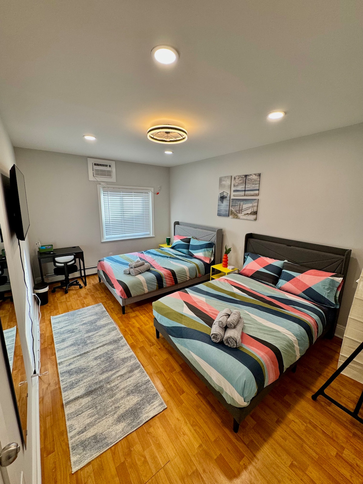 A private bedroom is presented featuring two spacious beds with colorful striped bedding. A small desk and chair are positioned in the corner, and a light-colored rug adds warmth to the hardwood floor. Natural light enters through the window, enhancing the comfortable ambiance.