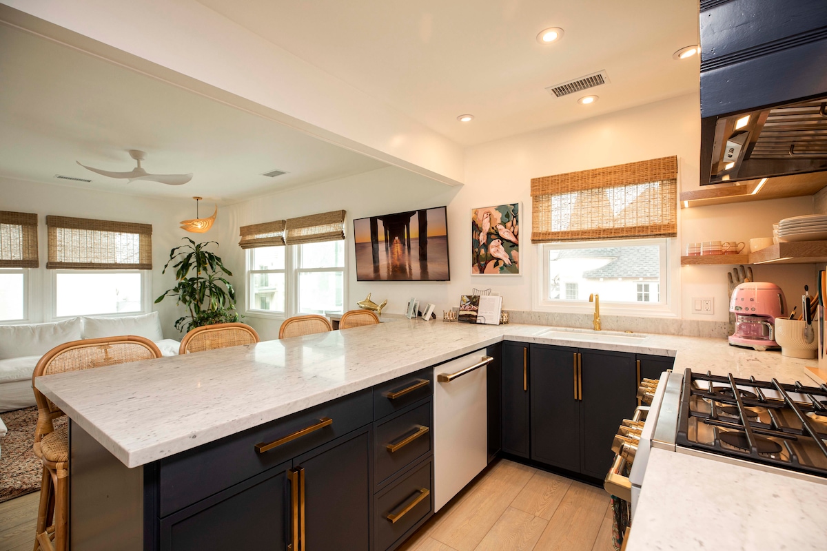 An open kitchen showcases sleek cabinetry in dark tones contrasted by a light marble countertop. Modern appliances, including a gas stove, are visible. Natural light filters in through windows dressed with bamboo shades, enhancing the airy feel of the space.