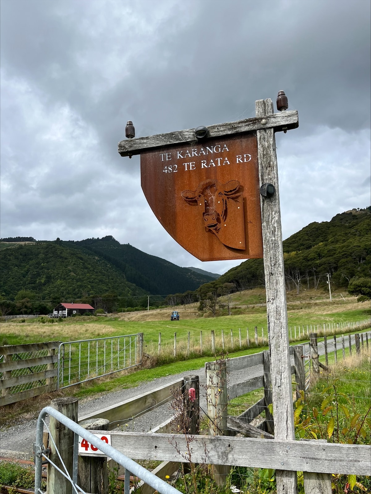 A rustic wooden signpost is positioned alongside a grassy path, partially obscured by a weathered metal panel. A lush landscape unfolds with rolling hills in the background, while a small building can be seen nestled among foliage. Overcast skies create a serene, muted atmosphere.