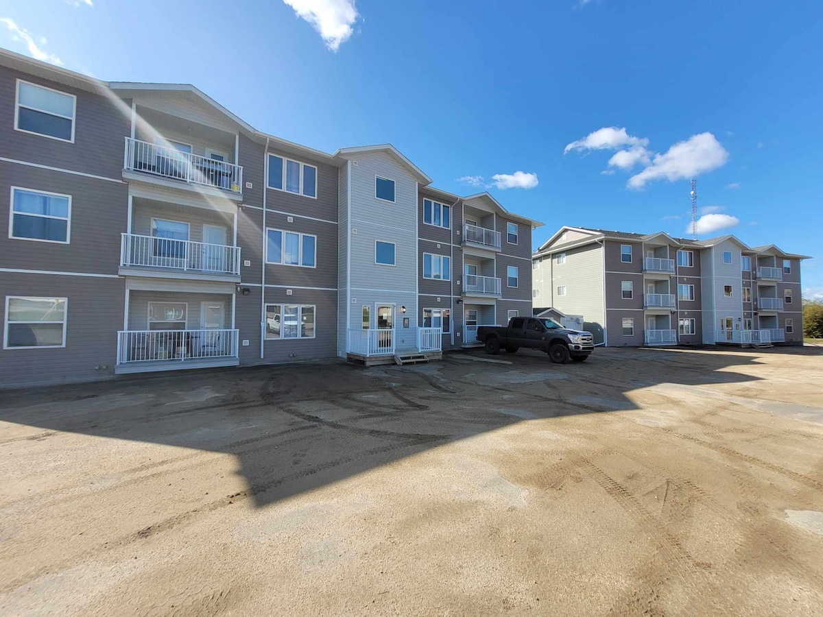 The exterior of a modern three-story apartment building is shown under a clear blue sky. The building features multiple balconies each with white railings. A spacious parking area accommodates vehicles, and neighboring buildings are visible in the background.