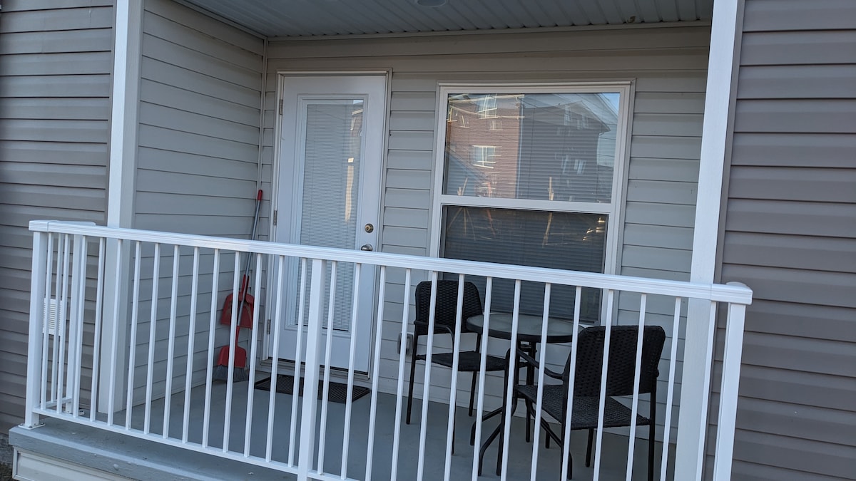 A small outdoor porch features a white railing and a neutral-colored floor. Two black wicker chairs are situated around a small table. A front door with a glass panel allows natural light to enter the interior space, while a window beside it provides additional brightness.